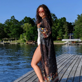 A woman standing on a wooden dock wearing a sheer black mosquito repelling getaway kimono with a floral design, in front of lake with boats and trees in the background.